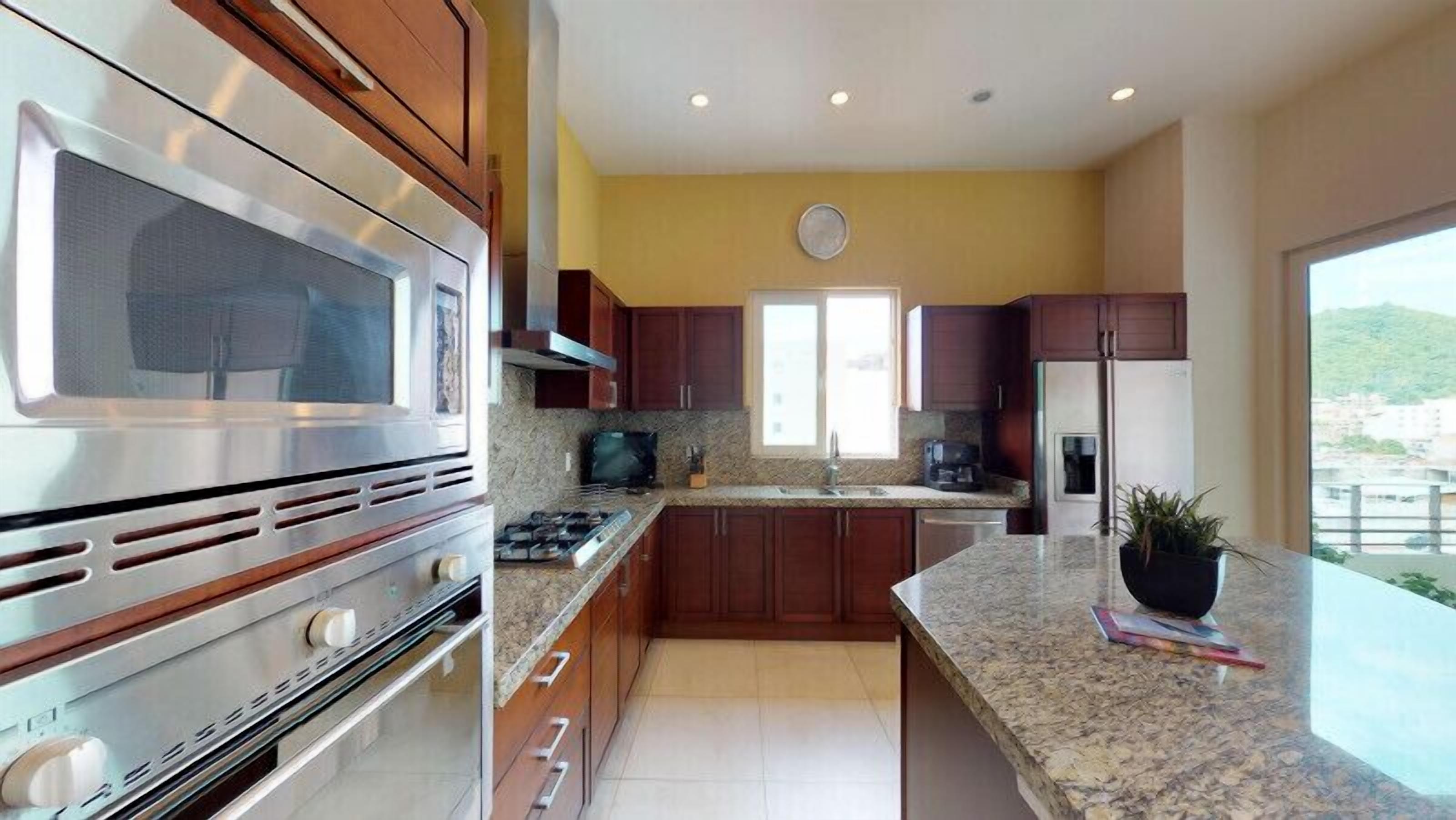 A modern kitchen with stainless steel appliances, wooden cabinets, granite countertops, and a large window offering a view.