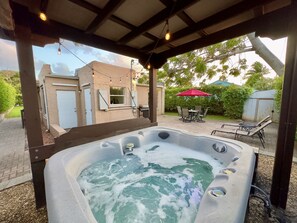 Jacuzzi bajo un porche cubierto con vista a una casa, una mesa con sillas y un paraguas rojo.