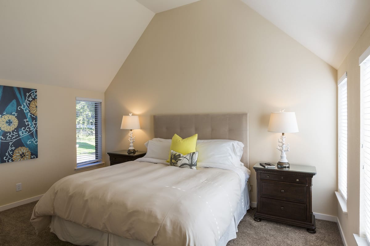 A bedroom with a large bed, two nightstands with lamps, a window with blinds, and a wall with a floral painting.
