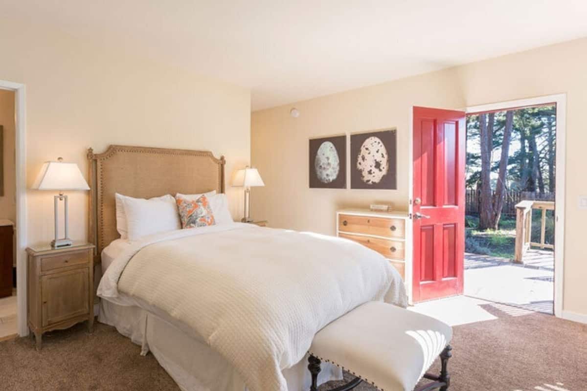 A bedroom with a large bed, bedside tables with lamps, a dresser, and a red door leading to a balcony.