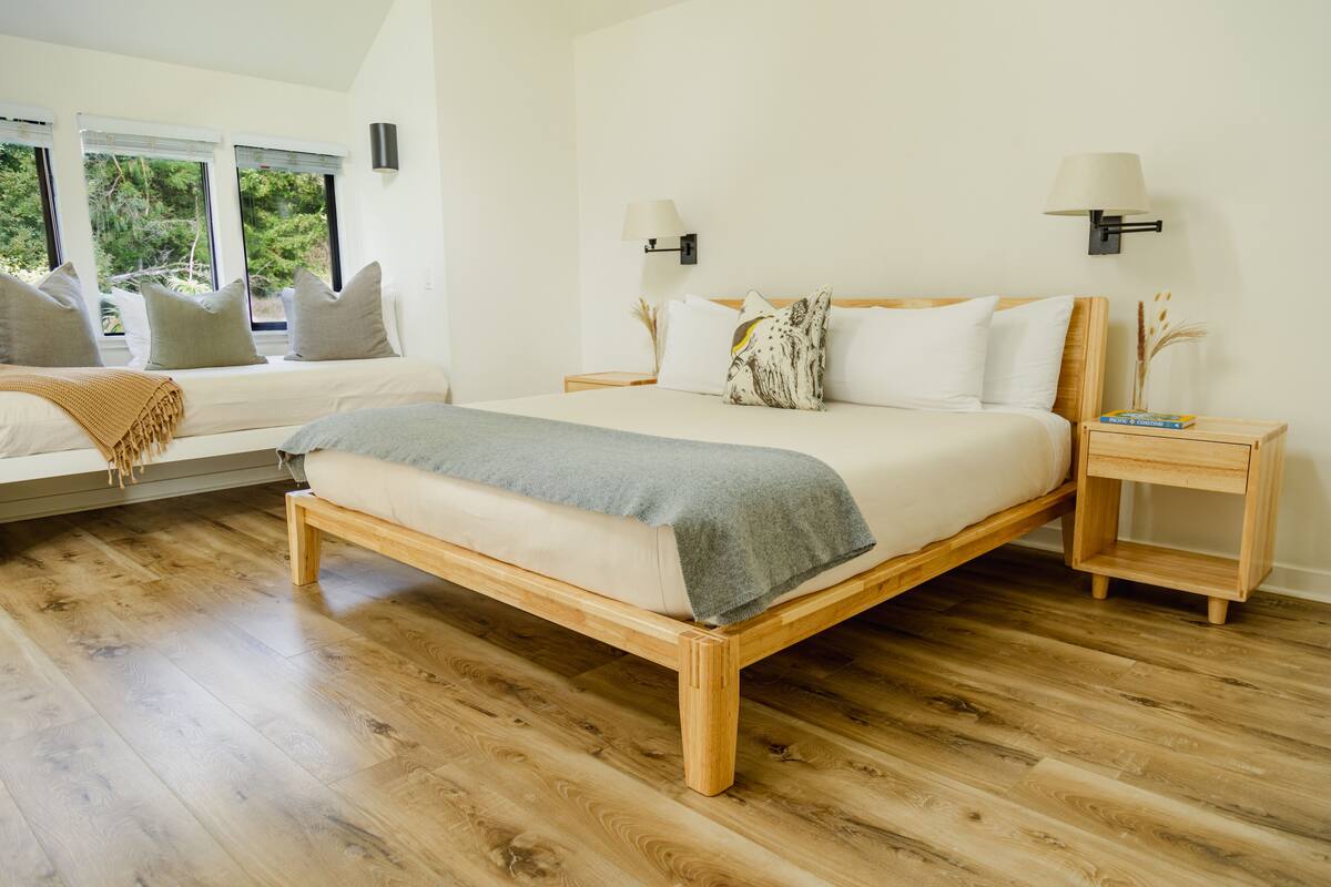 A modern bedroom with a wooden bed, two bedside tables, a window seat, and a view of greenery outside.