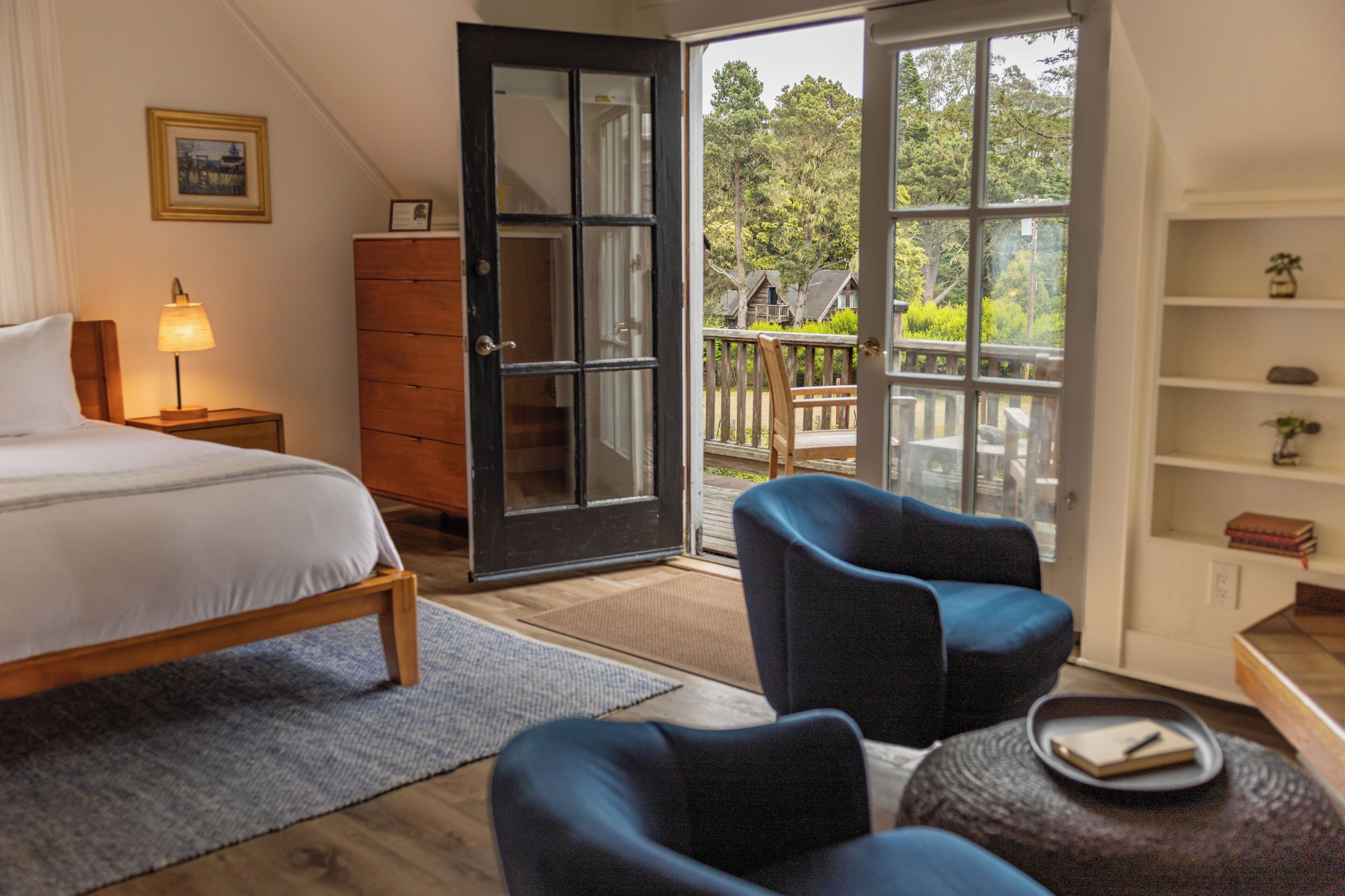 A bedroom with a bed, a desk, a chair, a dresser, and a balcony with a view of trees.