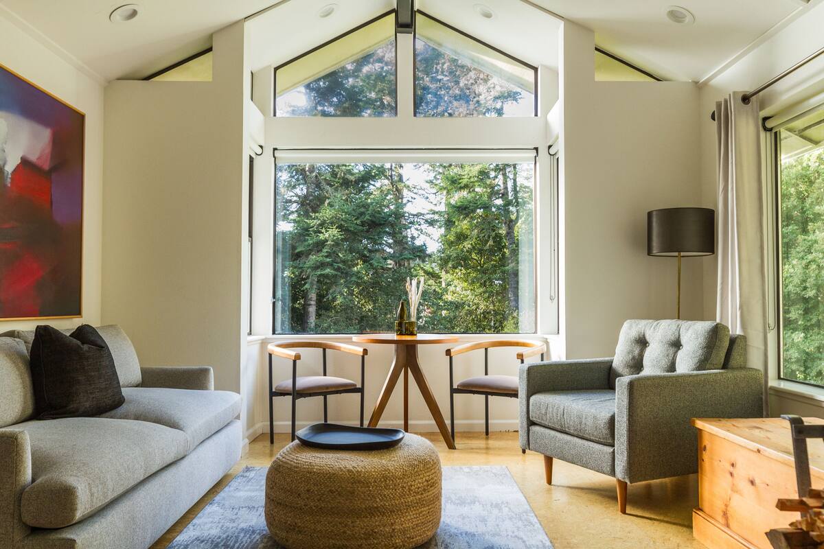 A modern living room with a high ceiling, a large window, a sofa, a chair, and a small table.