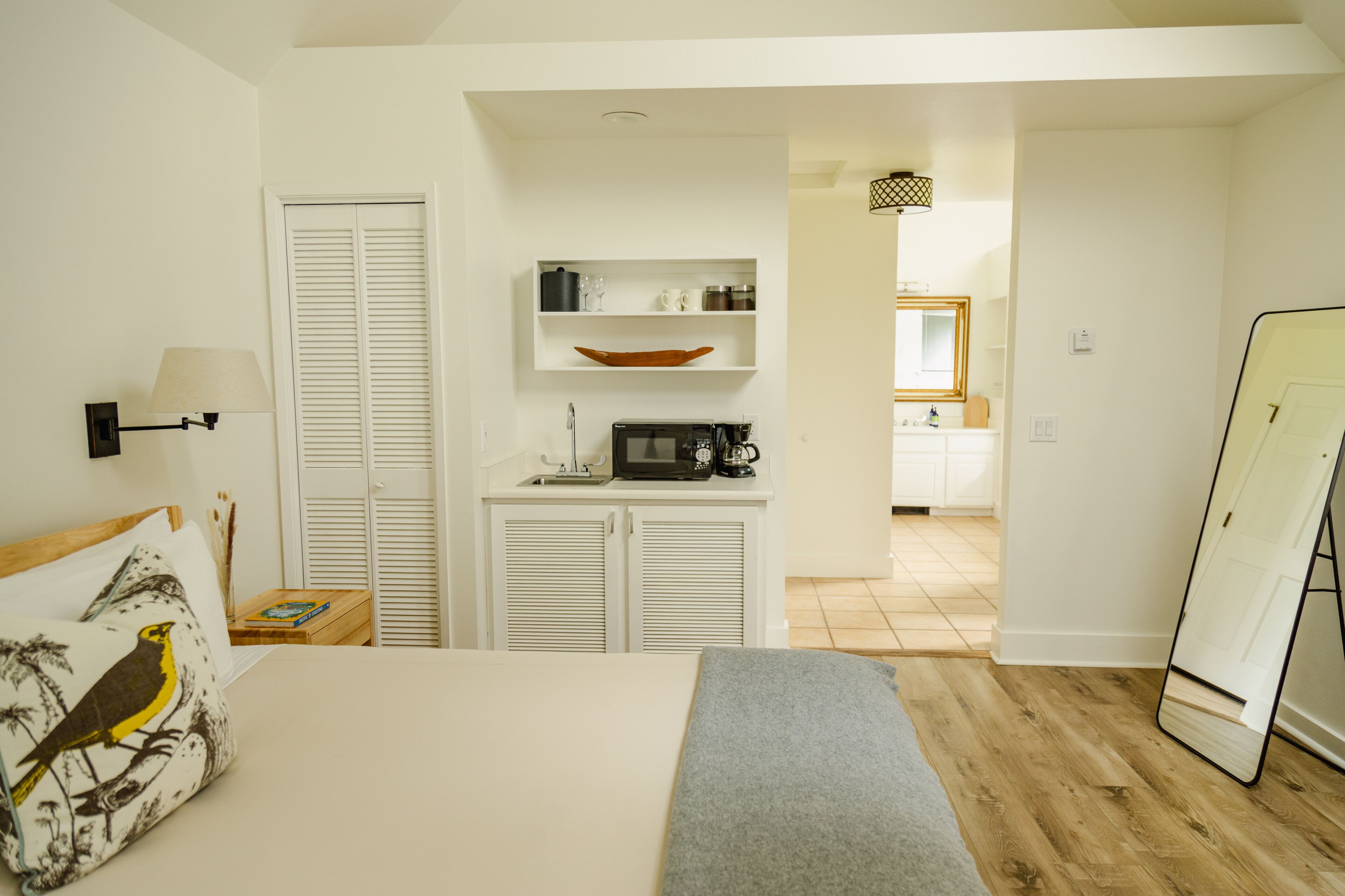 A bedroom with a bed, bedside table, lamp, and a view into the kitchen.