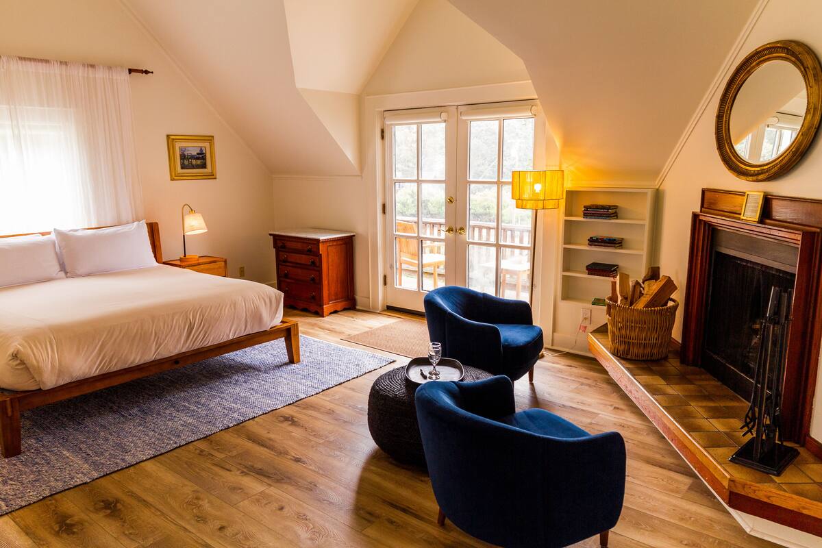A bedroom with a large bed, two chairs, a fireplace, and a bookshelf.