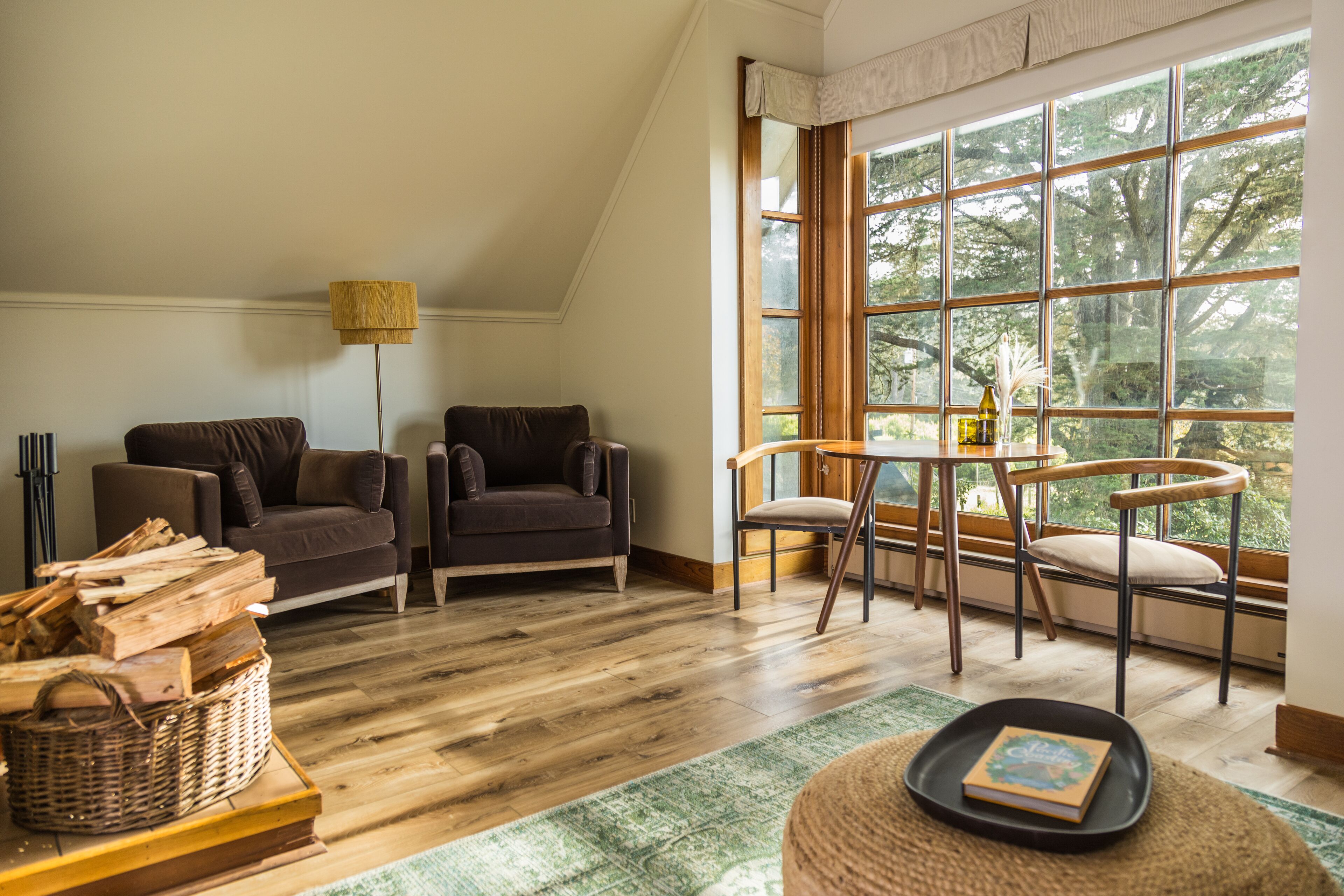 A cozy living room with a large window, wooden flooring, a sofa, a chair, and a small table with a book.