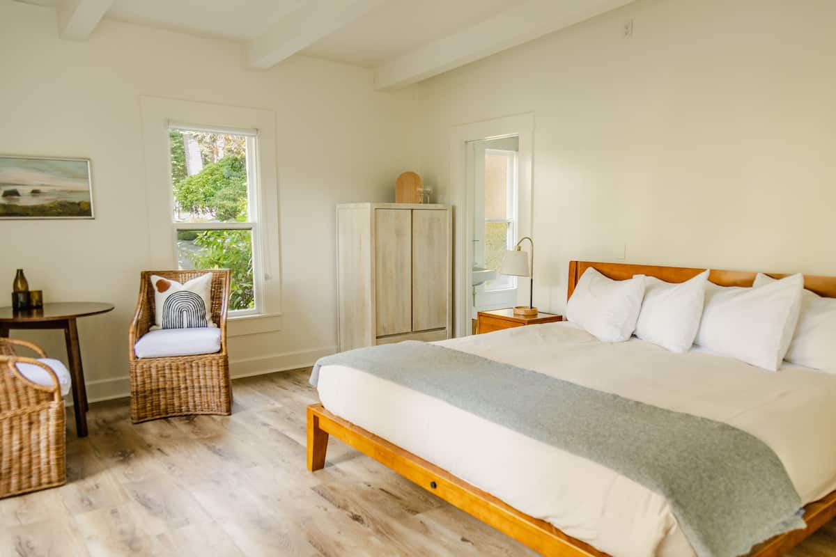 A bedroom with a large bed, a wicker chair, a wardrobe, and a small table.