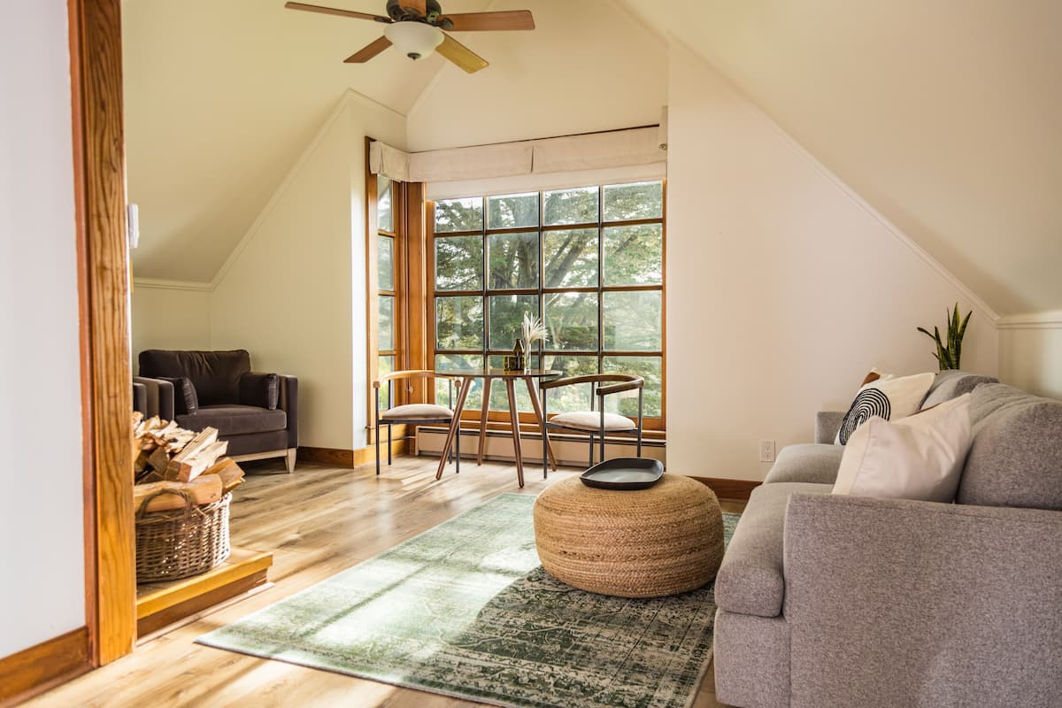 A spacious loft with a high ceiling, a large window offering a view of trees, a cozy seating area with a gray sofa and a round ottoman, and a wooden floor.