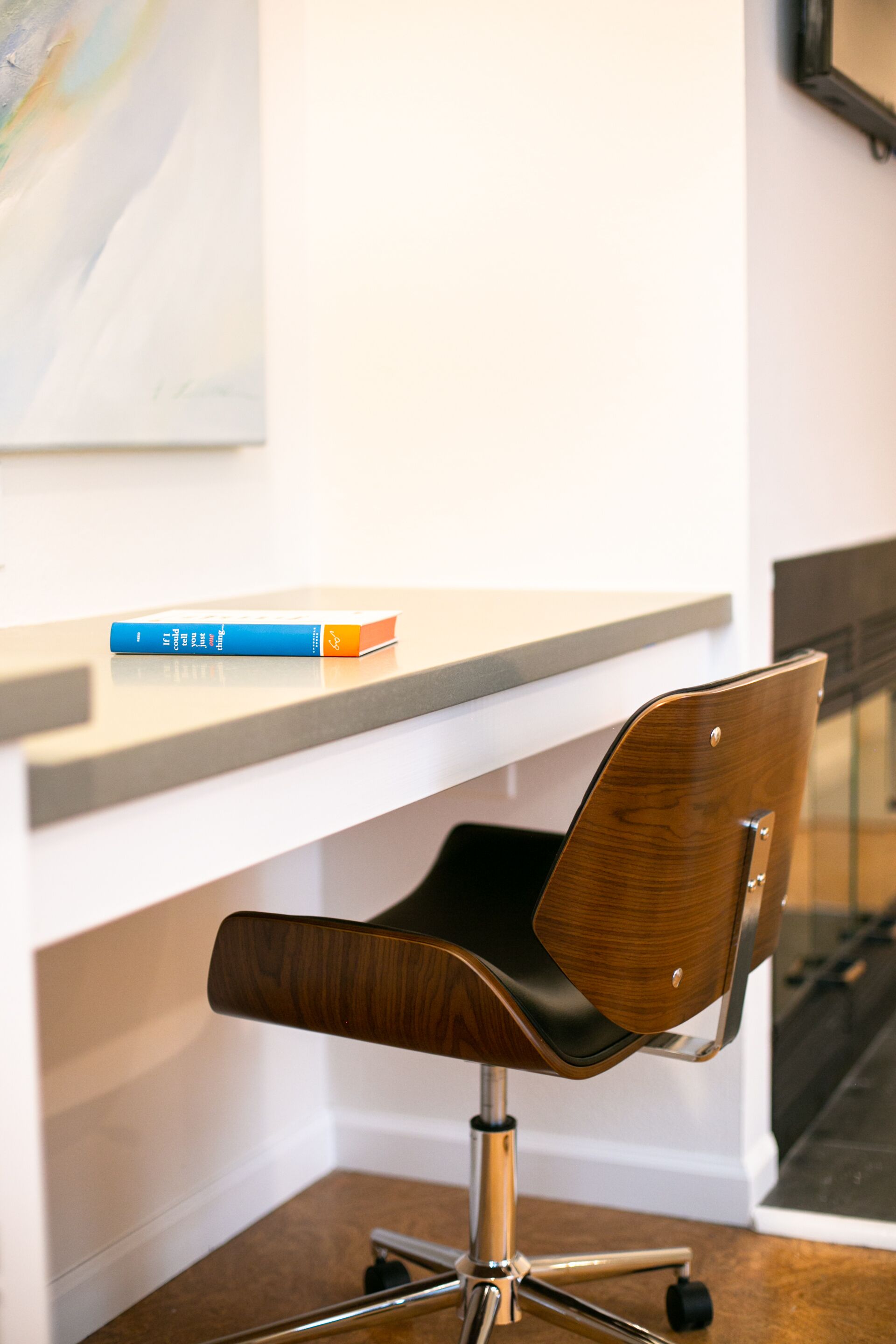 A modern office chair with a wooden backrest and metal frame, placed on a white desk with a book on it.