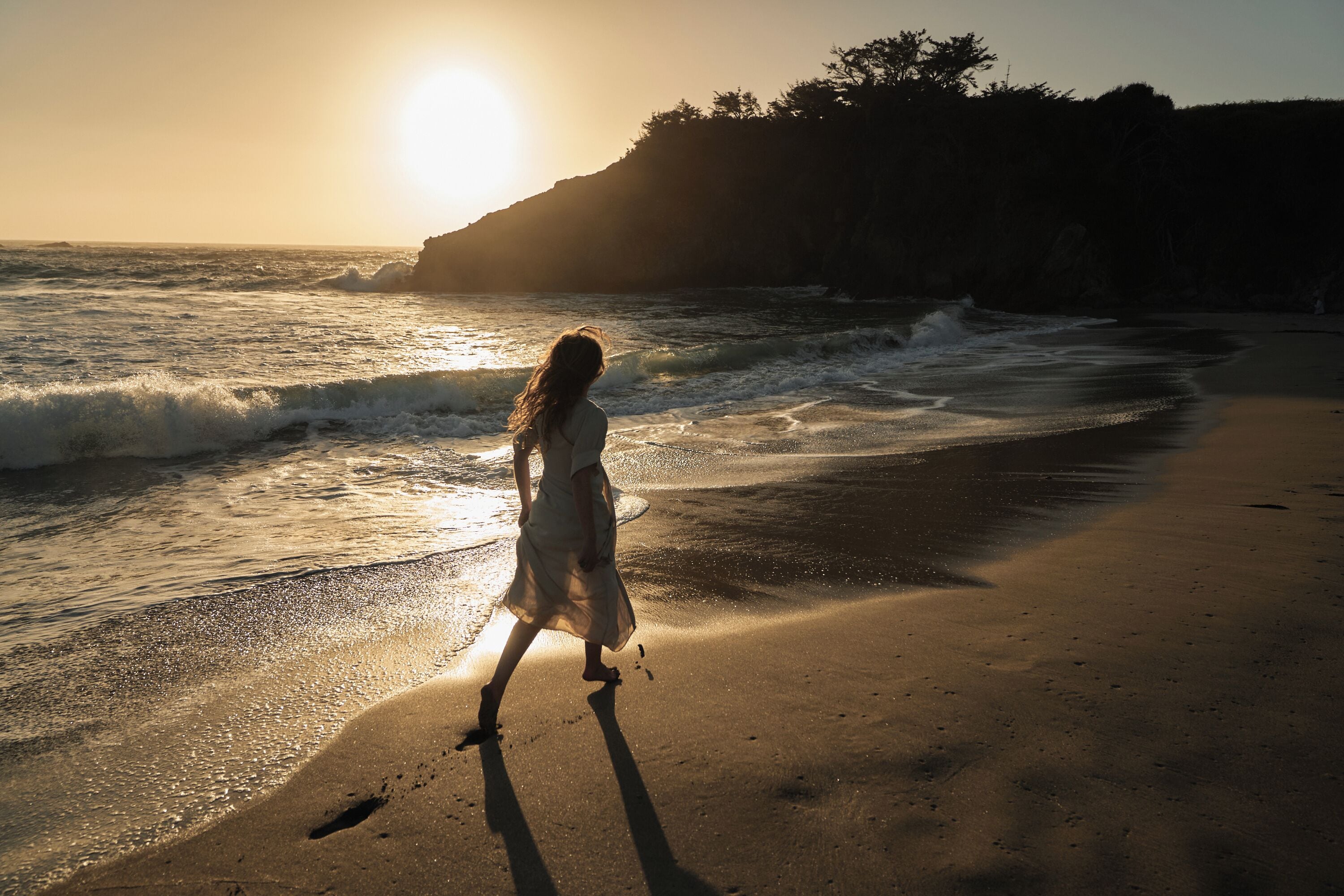 Escena de playa con una persona caminando por la orilla, olas rompiendo sobre la arena y el sol poniéndose al fondo.