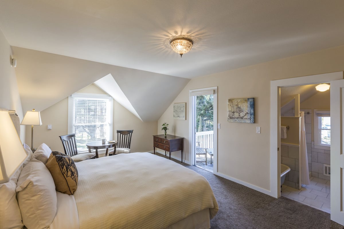 A bedroom with a large bed, a desk, and a chair. There is a window with blinds, a ceiling light, and a door leading to a balcony.