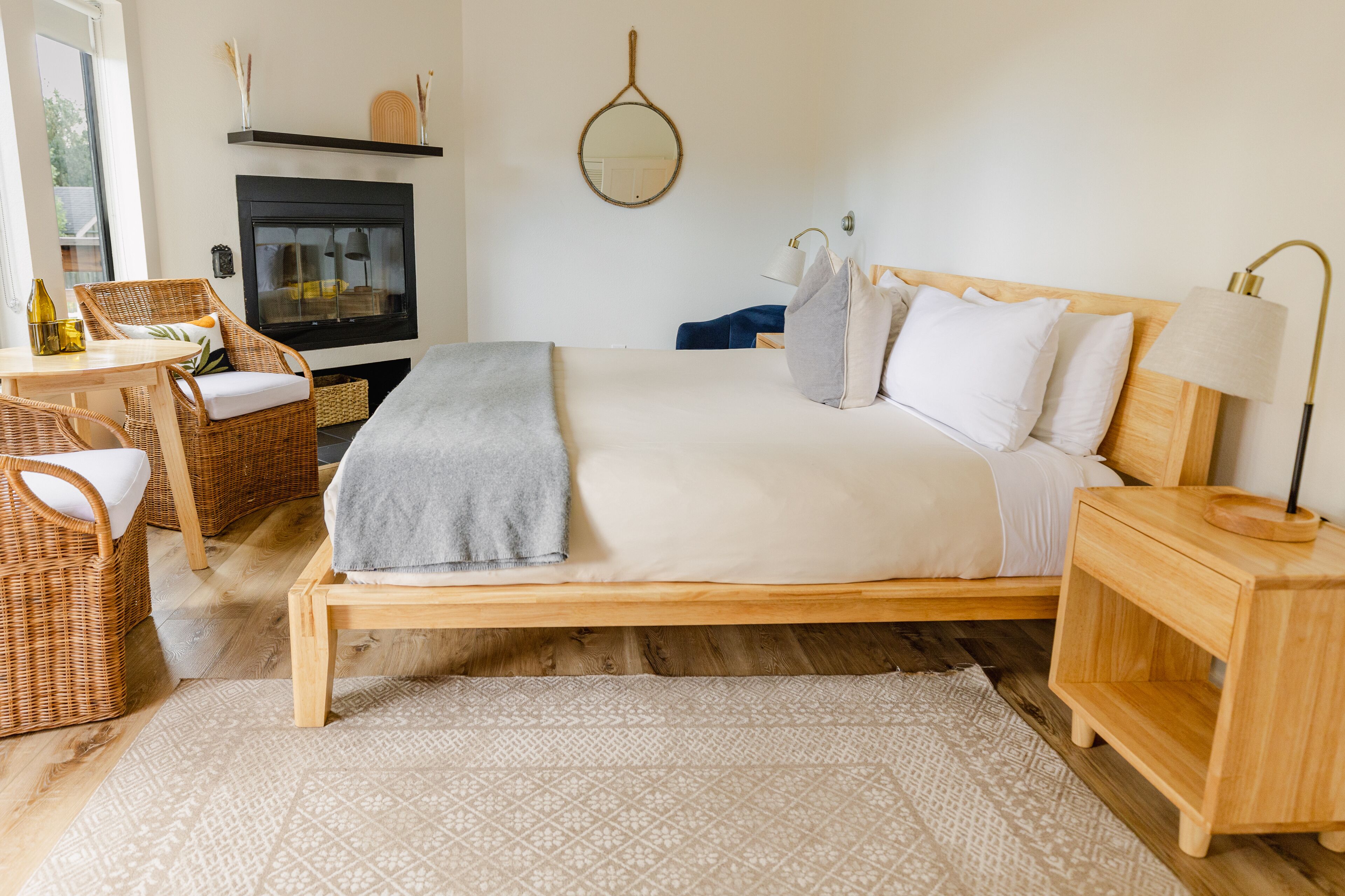 A bedroom with a wooden bed, a bedside table, a nightstand, a wicker chair, a small table with a lamp, and a fireplace.