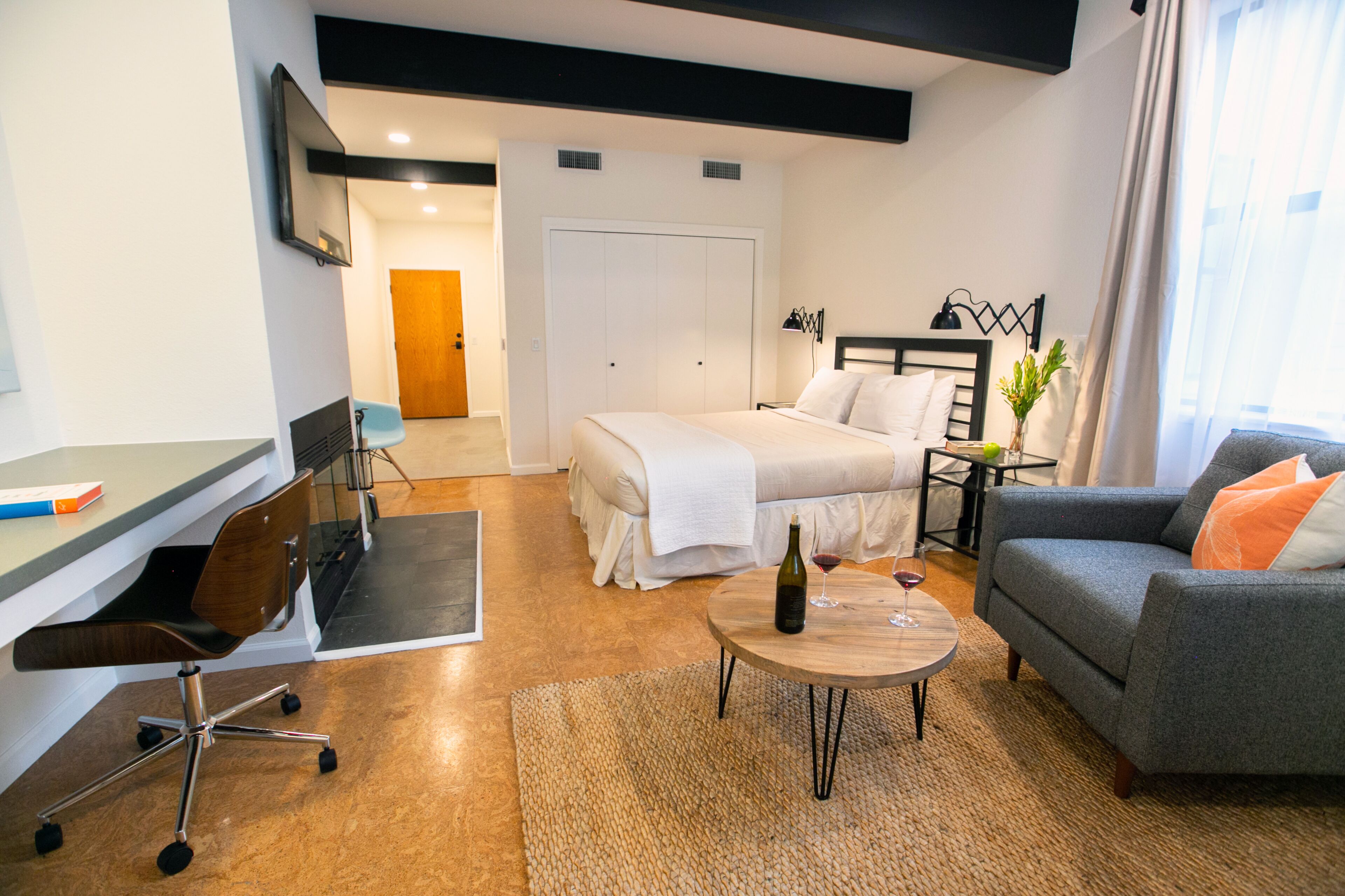 A modern hotel room with a bed, sofa, desk, and a small table with a bottle and glass.