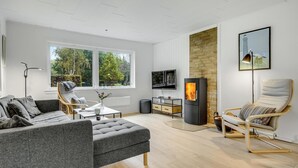 A modern living room with a grey sofa, a wooden floor, a TV, a fireplace, and a large window with a view of trees. - 3 bedroom cozy home in Nykøbing Sj (Nykøbing Sj)