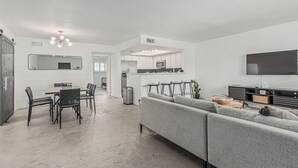 Una sala de estar moderna con un sofá gris, un comedor con una mesa y sillas de madera, una cocina con armarios blancos y un televisor montado en la pared.