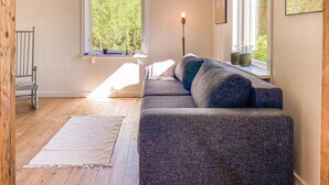 Uma sala de estar moderna com um sofá cinzento, piso de madeira, janela com vista para árvores e um abajur.
