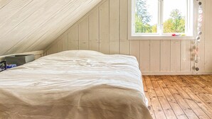 Quarto com teto inclinado, cama com colcha branca, janela com vista para árvores e piso de madeira.