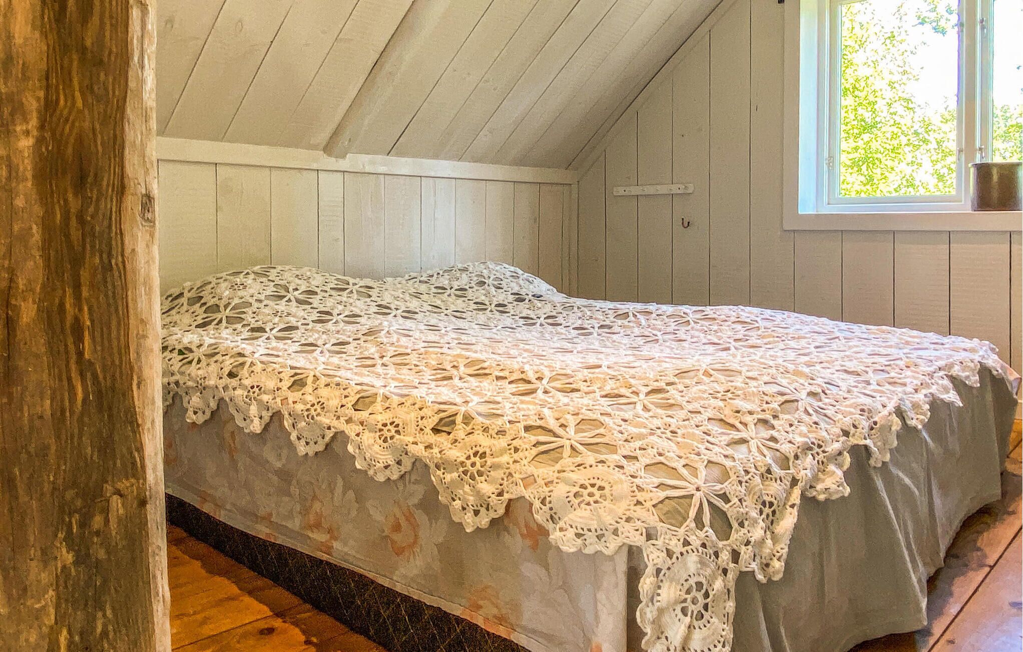 A room with a bed covered in a white lace bedspread, wooden walls, and a window with a view of greenery.