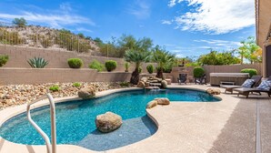 A swimming pool with a waterfall feature, surrounded by rocks and plants, with a patio area and outdoor furniture.