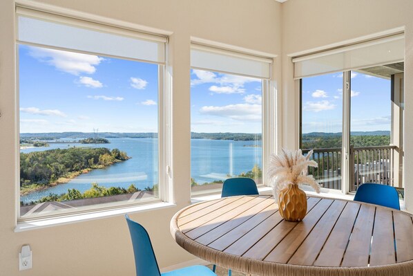 Mesa redonda de madera con sillas y un florero, con vista a un lago y un paisaje distante a través de amplias ventanas.