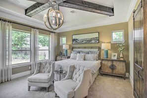 A bedroom with a large bed, two armchairs, a nightstand, a chandelier, and a view of the outdoors.
