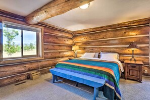 Un dormitorio de cabaña de madera con cama, banco, mesita de noche, lámpara y ventana grande.