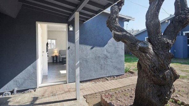 Une maison moderne dotée d’un porche abrité, d’un intérieur visible comprenant un coin repas, et d’un grand arbre au premier plan.
