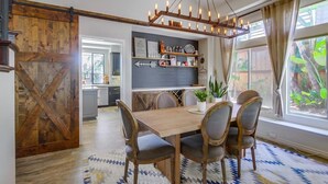 A dining area with a wooden table and chairs, a chandelier, and a view of the kitchen and garden through large windows. - Encinitas vacation home- 5 minutes from the beach (Encinitas)