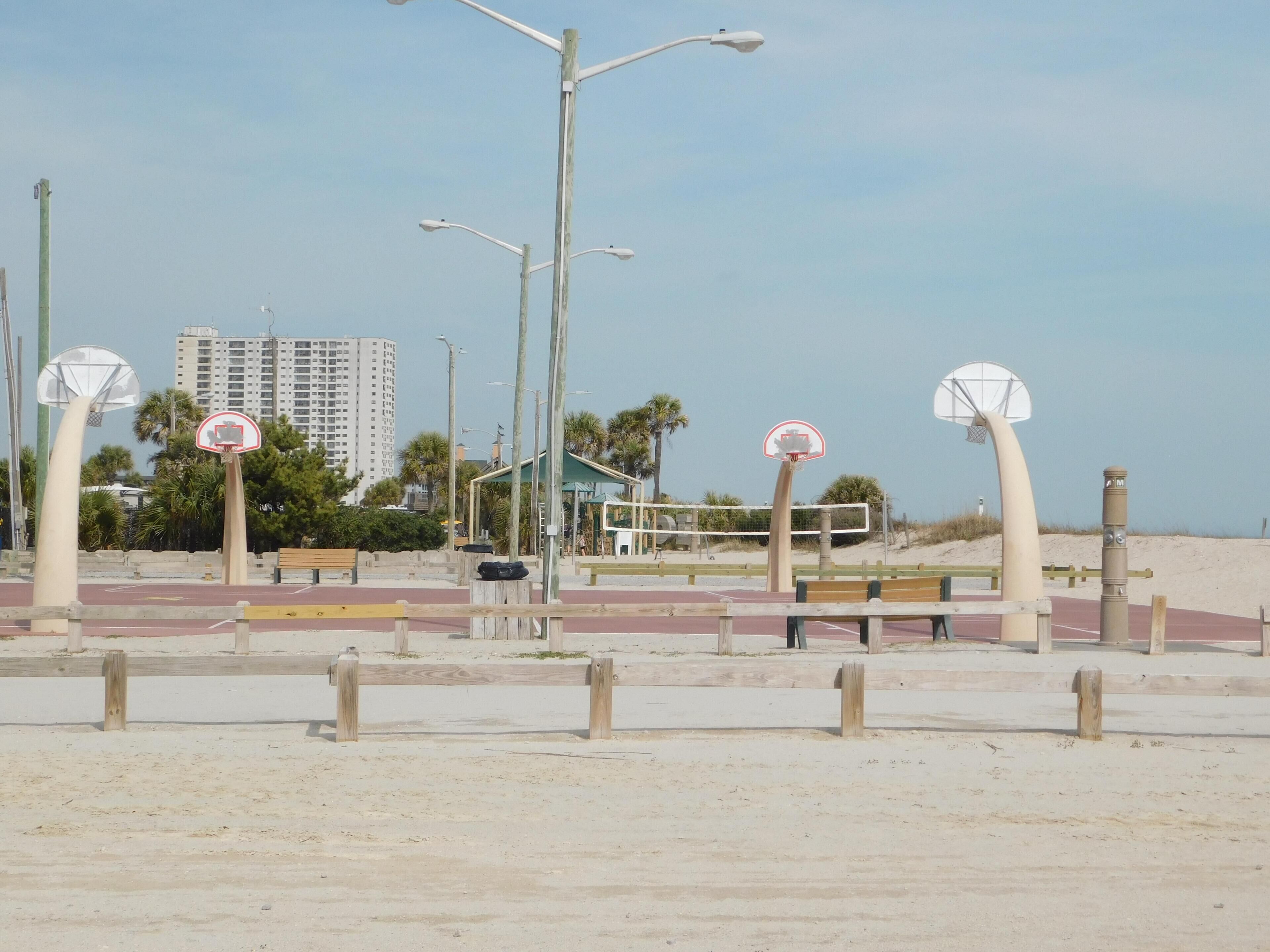 Una cancha de baloncesto a la orilla de la playa con un banco de madera, farolas y un edificio al fondo.