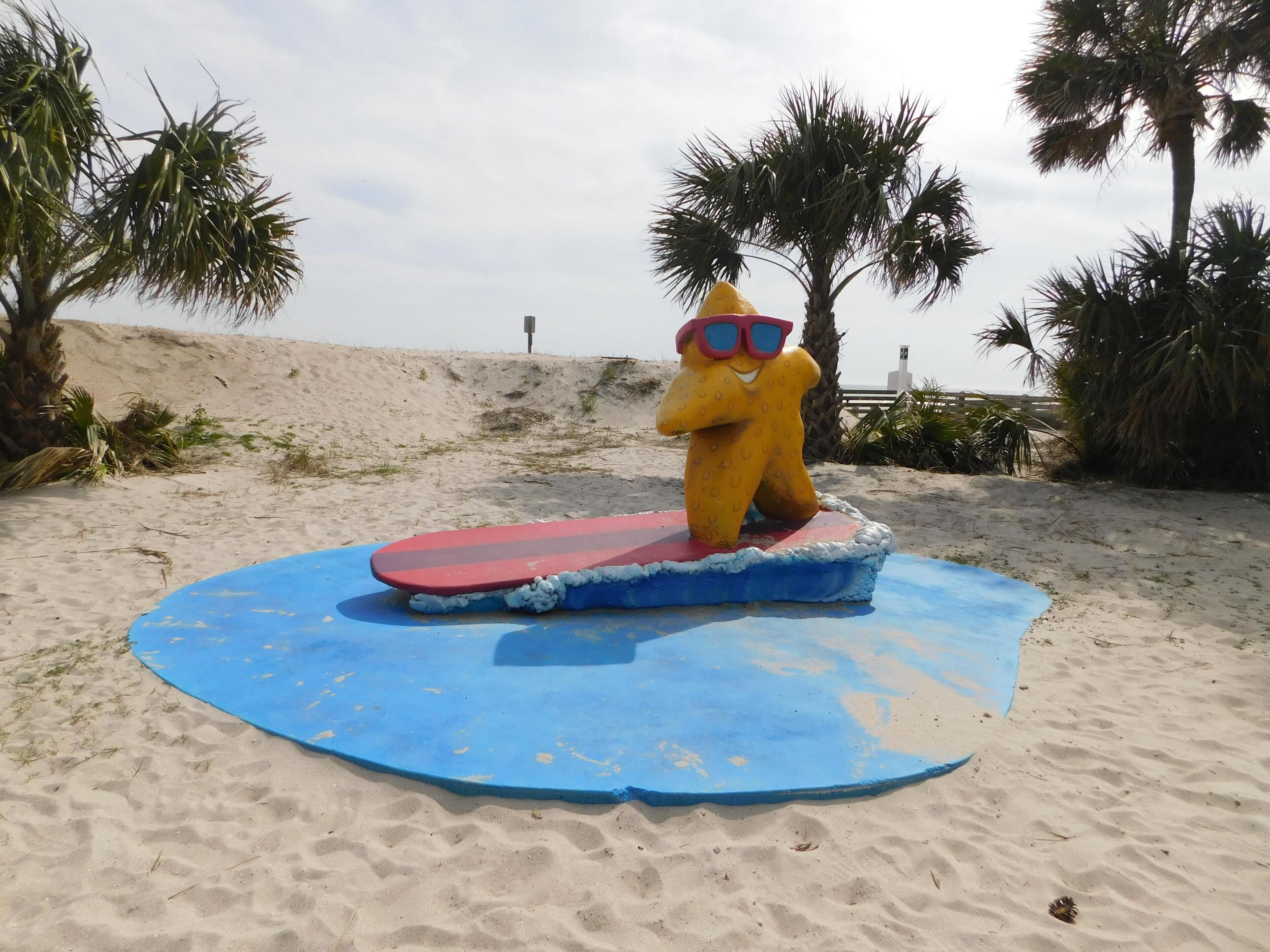 A beach scene with a yellow statue wearing sunglasses and a red surfboard.