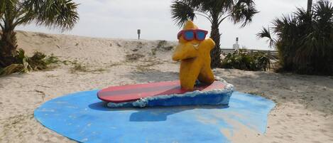 A beach scene with a yellow statue wearing sunglasses and a red surfboard.