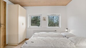 A bedroom with two beds, wooden ceiling, white walls, two windows with views of trees, and a radiator.