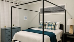 A four-poster bed with a white and teal bedspread, a bedside table with a lamp, a small bedside plant, and a dark blue dresser with drawers. - Stylish 3BR Home Sleeps 8 w/ Grill – Near CrossPlex & Downtown (Birmingham)