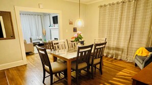 A dining area with a wooden table and chairs, a kitchen in the background, and a living area with a sofa and a lamp. - Stylish 3BR Home Sleeps 8 w/ Grill – Near CrossPlex & Downtown (Birmingham)