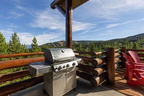 A barbecue grill on an outdoor deck with a wooden railing and red chairs, overlooking a mountainous landscape.