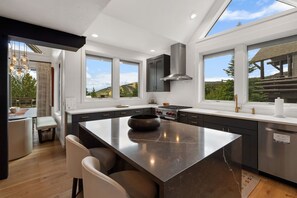 A modern kitchen with a large island, dark cabinetry, and a view of a forested landscape through large windows.