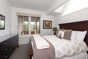 A bedroom with a large bed, a dresser, a window with blinds, a painting on the wall, and a skylight.