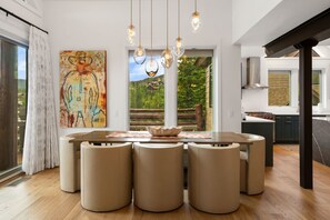 A modern dining area with a round table, six cushioned stools, pendant lighting, and a large abstract painting on the wall. - Ski-In/Ski-Out 3 Bedroom Moonlight Basin (Big Sky)