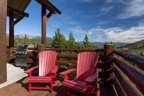 A wooden deck with two red Adirondack chairs, a grill, and a view of mountains.