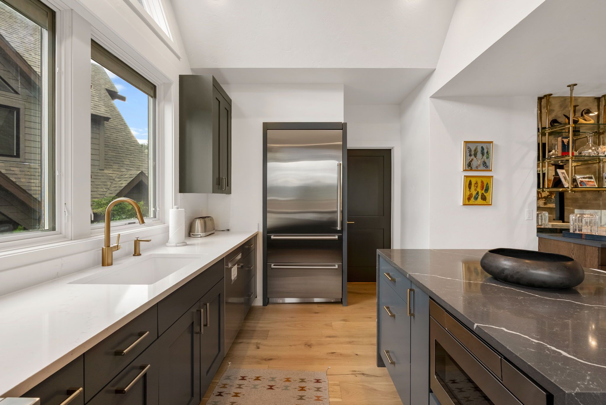 A modern kitchen with a stainless steel refrigerator, a large window, and a wooden floor.