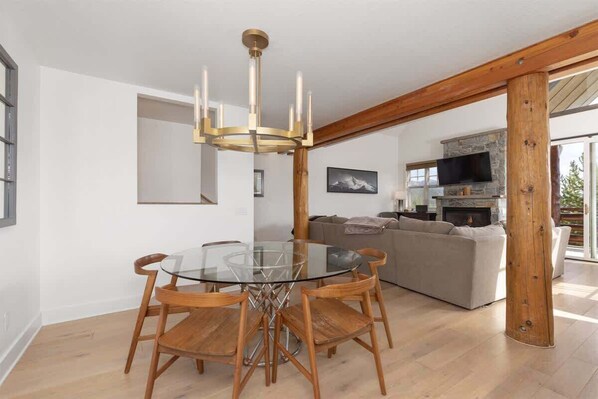 A modern living room with a glass dining table, wooden chairs, a stone fireplace, and a large window.