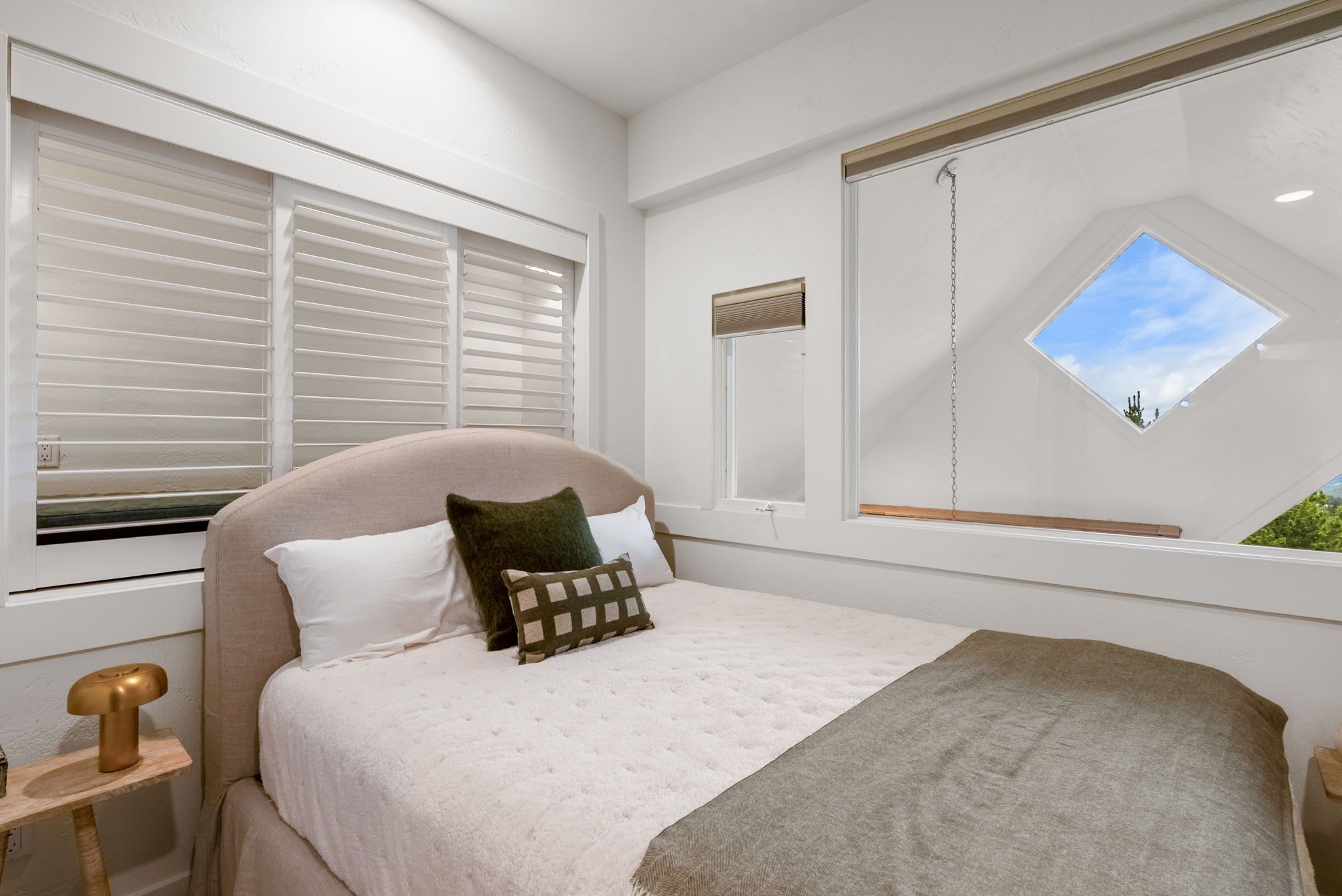 A bedroom with a bed, a nightstand, a window with blinds, a mirror, and a skylight.