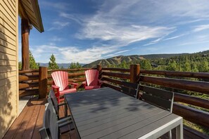 A wooden deck with a table and two Adirondack chairs, overlooking a forested landscape.