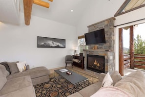 A modern living room with a stone fireplace, a flat-screen TV, and a large window overlooking a deck.
