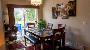 Un coin repas comprenant une table en bois, quatre chaises, un banc et un lustre. On y trouve une grande fenêtre avec des rideaux, une plante sur la table et des œuvres d’art accrochées au mur.