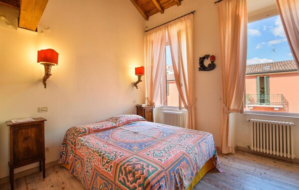 Een slaapkamer met een bed, twee wandlampen, een klein houten nachtkastje, een radiator en een raam met gordijnen.