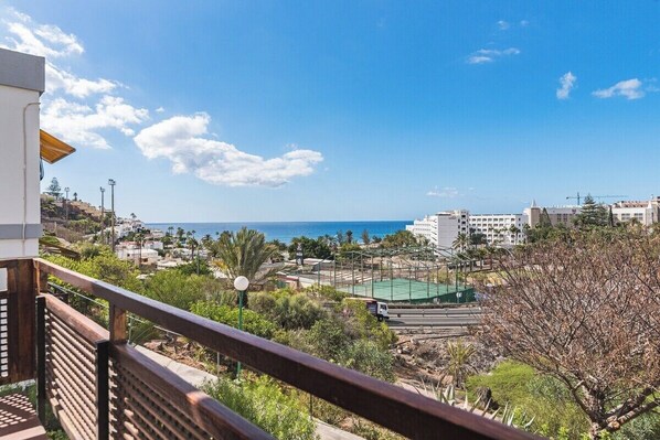 Uma varanda com vista para uma cidade costeira, incluindo edifícios, uma estrada e quadra esportiva verde.