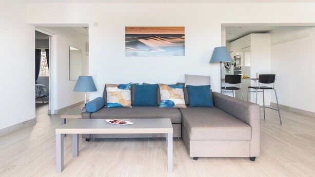 A modern living room with a grey sofa, a wooden coffee table, and a painting above the sofa. There is a dining area with a table and chairs in the background. - Home2Book Stunning Apartment Rocas Rojas (Maspalomas)
