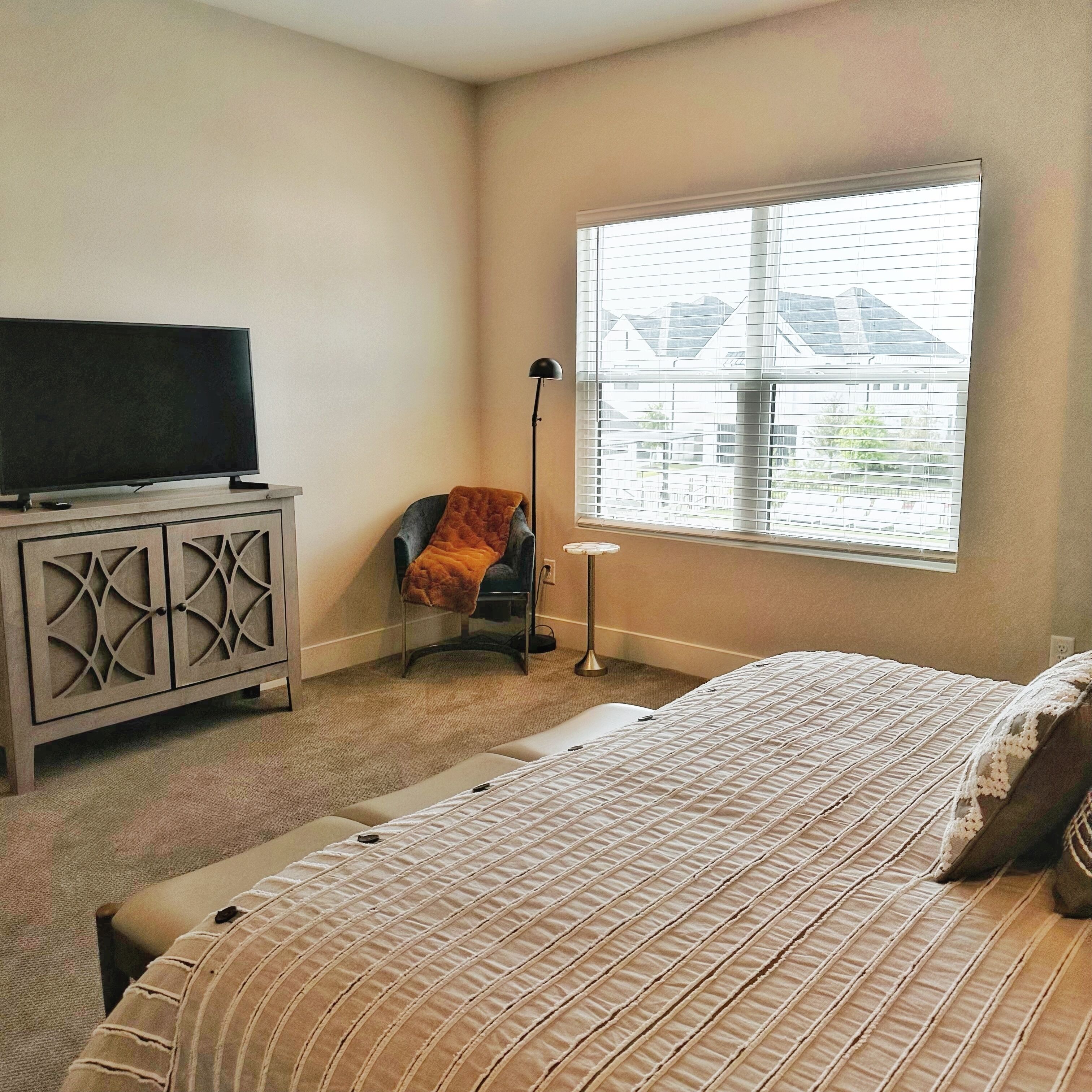 A bedroom with a bed, a TV on a cabinet, a chair, a floor lamp, and a window with a view of houses.
