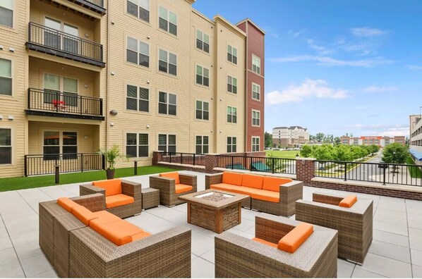 A rooftop patio with wicker furniture and a fire pit, surrounded by a pool and a view of a cityscape.